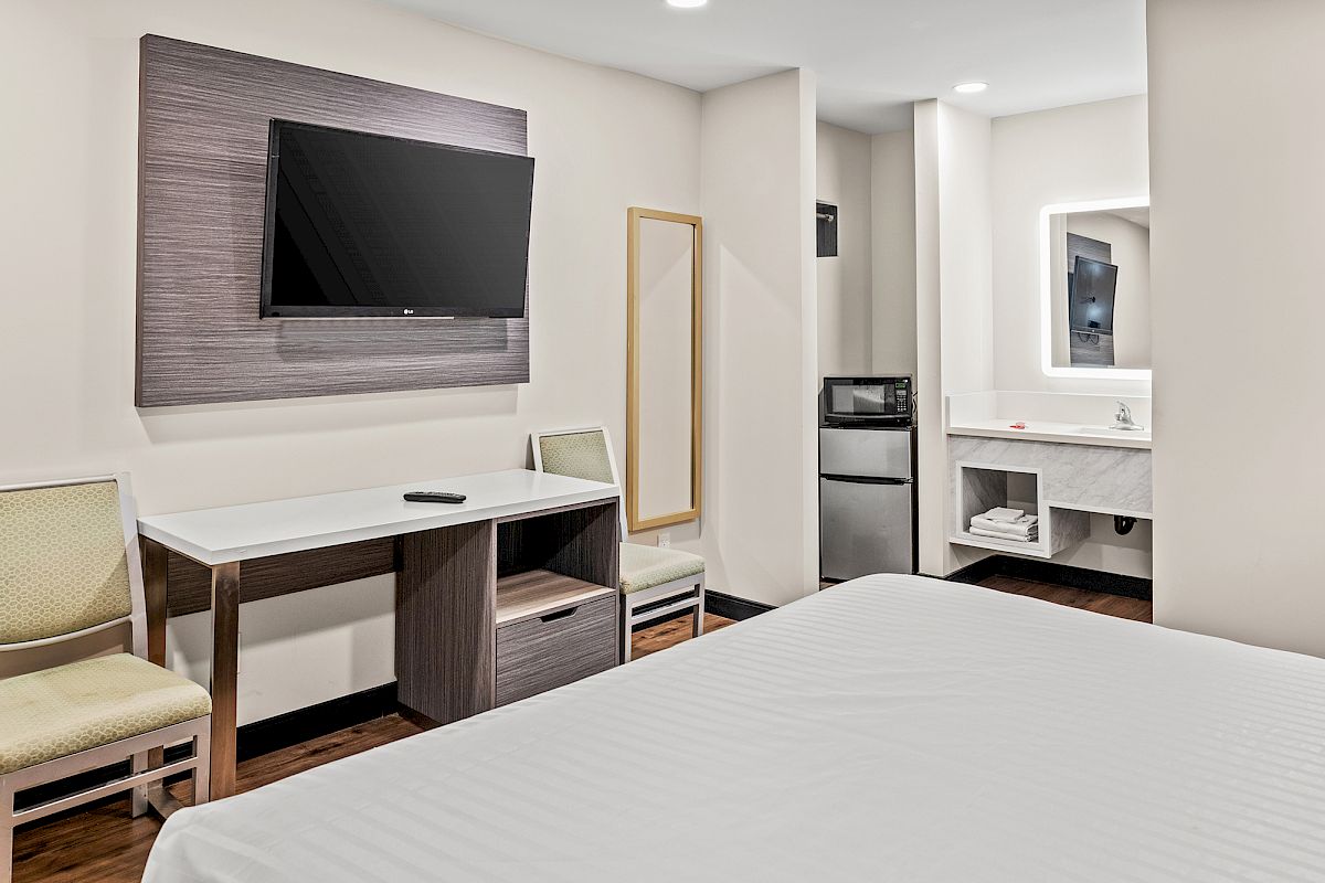 A hotel room with a bed, desk, chair, wall-mounted TV, fridge, microwave, mirror, and minimal decor, creating a cozy ambiance.