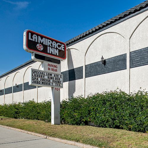 The image shows a sign for "La Mirage Inn" with a special offer, next to a white building with black accents and hedges in front.