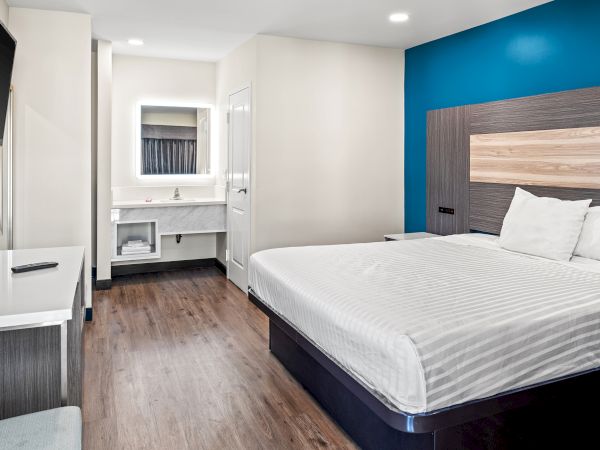 The image shows a modern hotel room with a large bed, blue accent wall, desk, wall-mounted TV, and a separate bathroom sink area.
