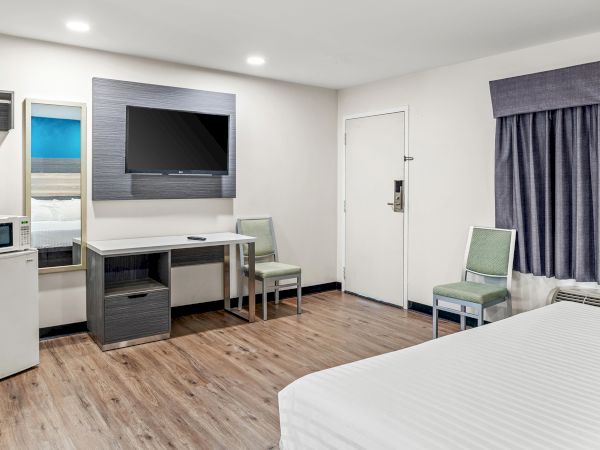 The image shows a hotel room with a bed, TV, desk, microwave, mini-fridge, and chairs, featuring wooden flooring and simple decor.
