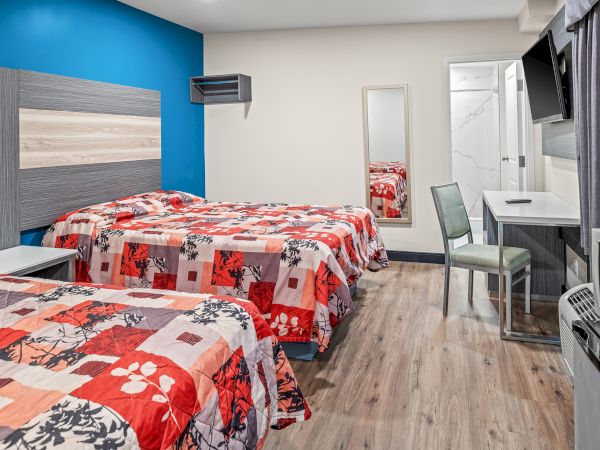 The image shows a modern hotel room with two beds, a desk, chair, TV, and bright decor. The floor has wood-style flooring.