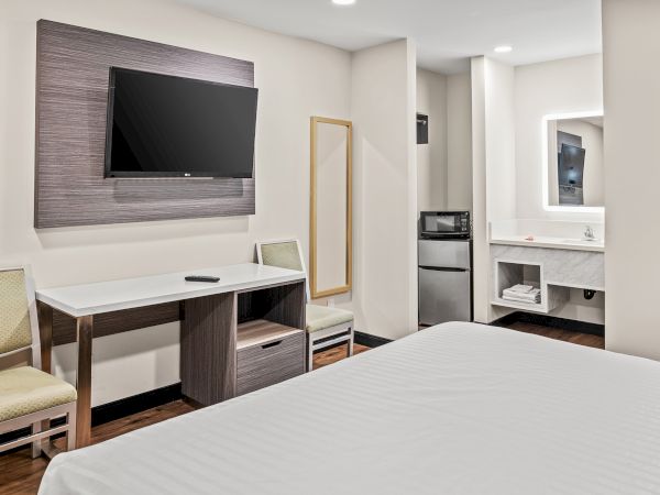 The image shows a hotel room with a TV, desk, chair, mirror, microwave, and mini-fridge. The room has neutral colors and modern decor.