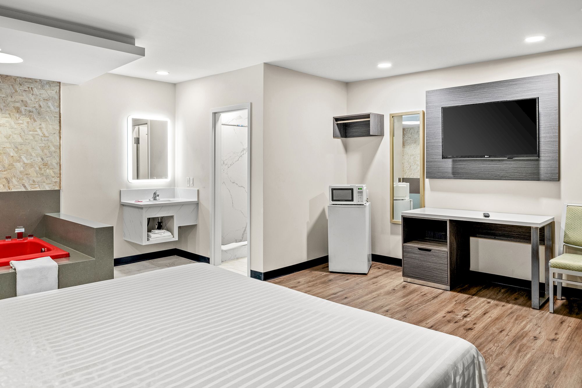 A wide-angle view of a hotel room with a king-sized bed, desk, microwave, TV, and red bathtub.