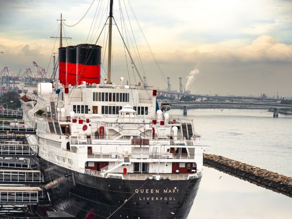 A large, historic ship docked at a harbor, surrounded by calm waters and a rock barrier. The ship has a red and black smokestack.