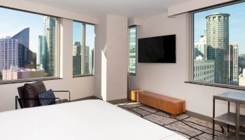 A modern hotel room with a bed, a chair, a wall-mounted TV, and large windows showing a cityscape with skyscrapers.