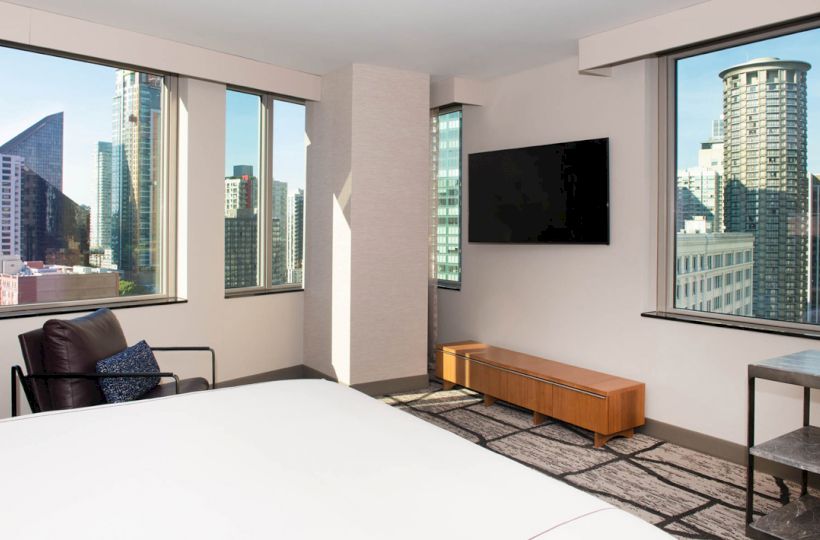 A modern hotel room with a bed, a chair, a wall-mounted TV, and large windows showing a cityscape with skyscrapers.