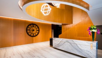 A modern lobby with a large clock on a wooden wall, white marble reception desk, spiral staircase, and decorative lighting.