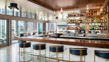 A modern bar with high stools, overhead lights, and shelves stocked with bottles. Large windows provide views of a cityscape outside.