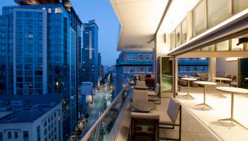 A modern rooftop terrace with seating overlooks a cityscape of tall buildings at dusk, with lights illuminating the street below.