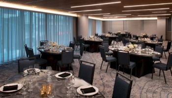 The image shows an elegant banquet hall set with round tables, dark chairs, tableware, and decorative elements, under modern lighting.