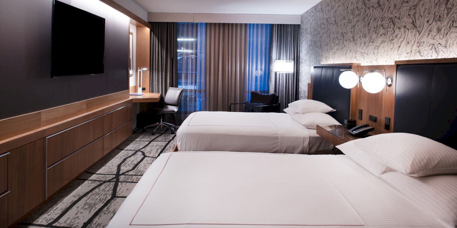A modern hotel room with two beds, a desk, a TV, and a window with curtains. The room has contemporary decor and good lighting.