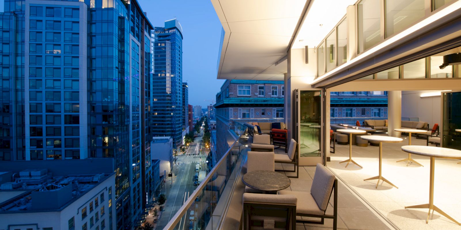 The image shows a modern rooftop terrace with tables and chairs, overlooking a cityscape with tall buildings at dusk.