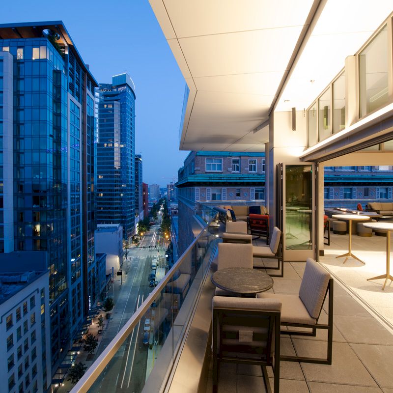 A modern urban balcony overlooks a city street, adjacent to tall buildings, featuring outdoor seating and warm interior lighting.