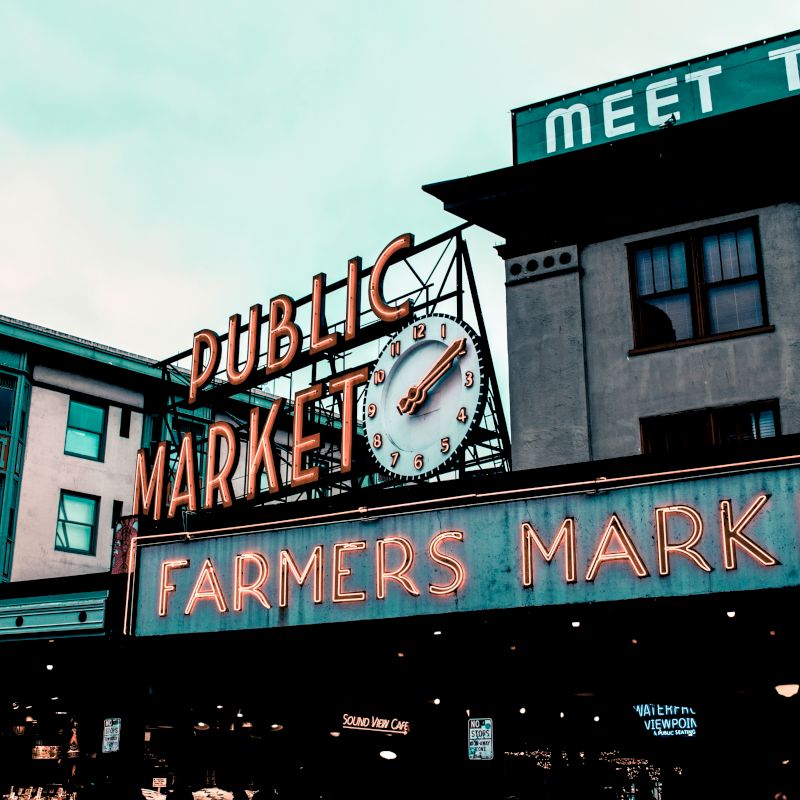 The image shows a neon sign for a public farmers market with a clock, set against a backdrop of buildings.