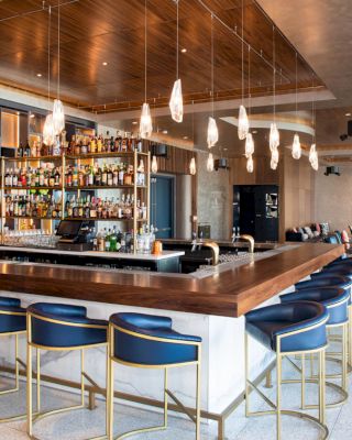 A modern bar with blue stools, a well-stocked shelf, and elegant lighting. The space is open with additional seating in the background.