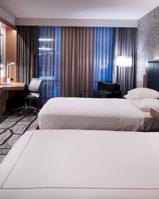 The image shows a modern hotel room with two beds, a large TV, a desk, and patterned carpet, featuring stylish lighting and wall design.
