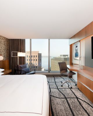 The image shows a modern hotel room with a bed, large TV, desk, chair, and window with a city view. The decor is contemporary and minimalistic.