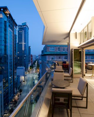 A modern cityscape at dusk with tall buildings and a stylish, open balcony featuring chairs and tables overlooking the street below.
