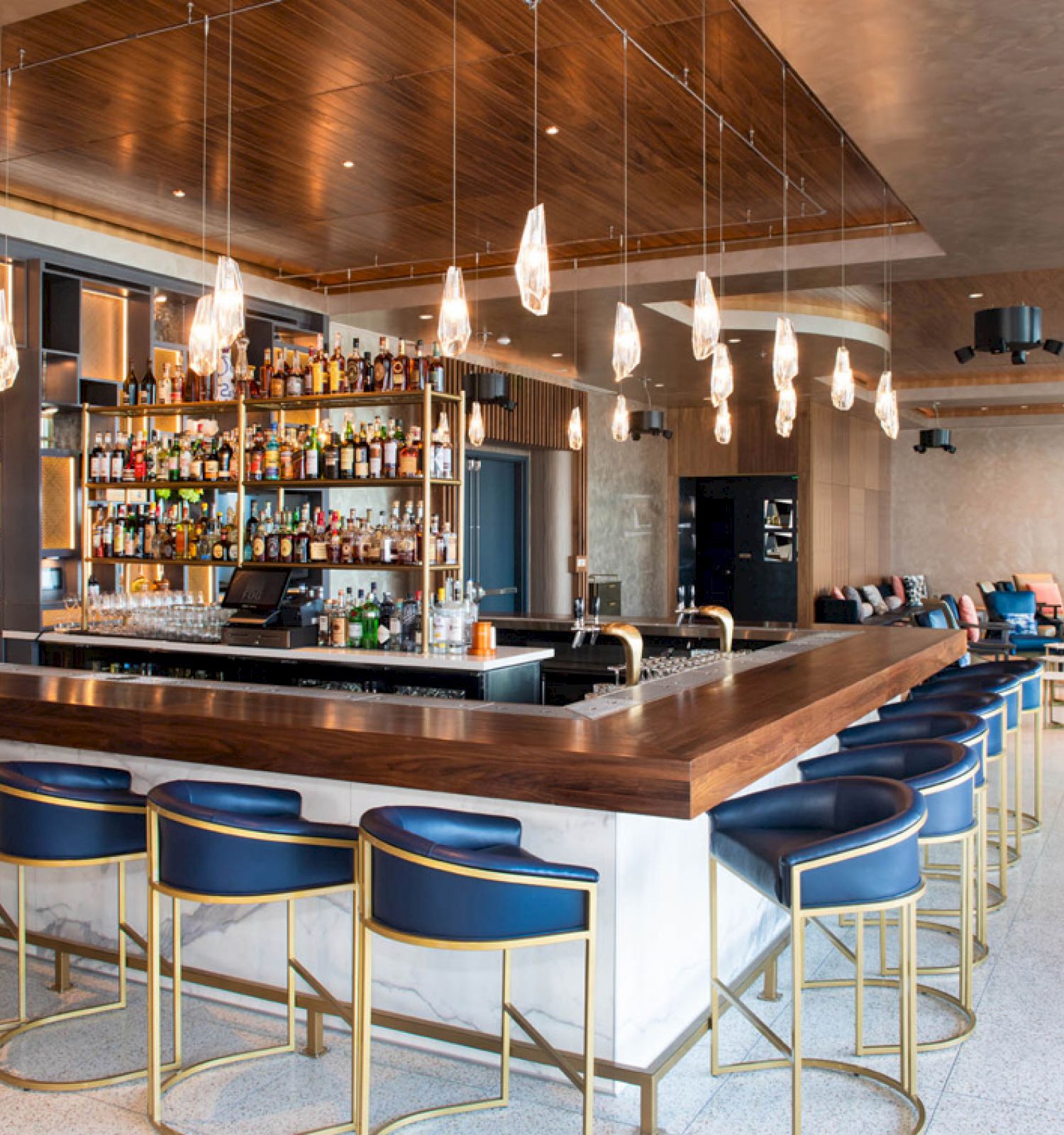 A modern bar with blue chairs, hanging lights, and a well-stocked liquor shelf, located in a spacious lounge area with seating.