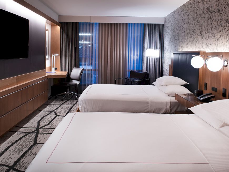 The image shows a modern hotel room with two double beds, a large TV, a work desk, and stylish decor, creating a cozy atmosphere.