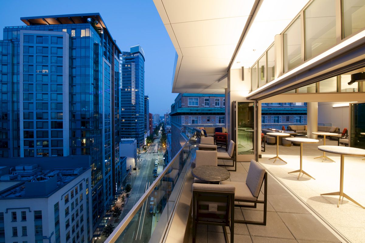 A modern cityscape with tall glass buildings is viewed from a stylish terrace with tables and chairs at dusk.