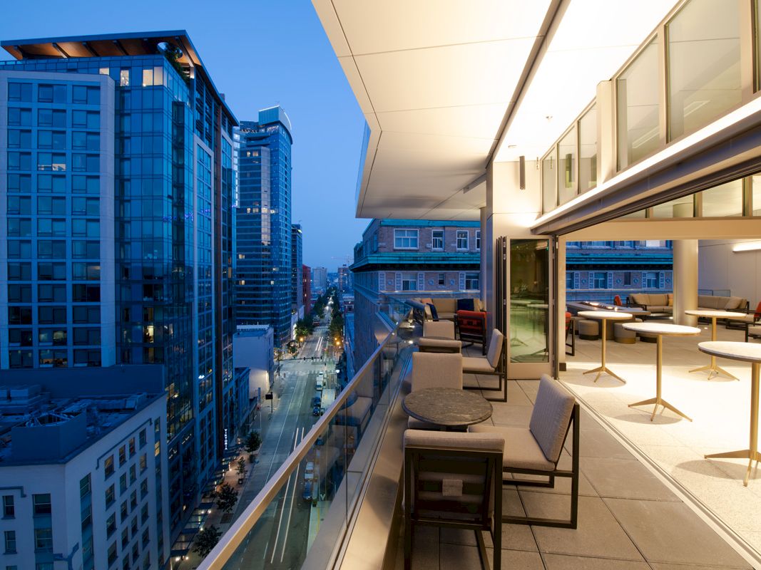 A modern cityscape with tall glass buildings is viewed from a stylish terrace with tables and chairs at dusk.