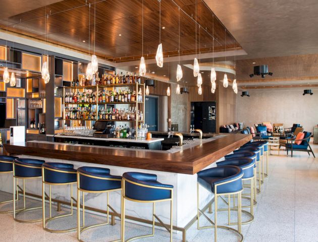 A modern bar with a large selection of bottles, stylish lighting, and seating. The interior is sleek with a cozy seating area nearby.