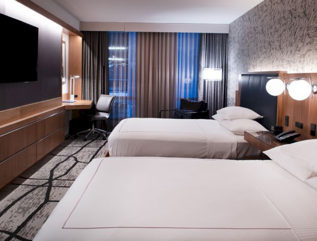 A modern hotel room with two beds, a wall-mounted TV, desk, chair, and stylish decor, featuring curtains and lamps for ambiance.