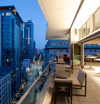 A modern urban balcony is pictured at dusk, overlooking a city street with tall buildings. The balcony has seating and tables.