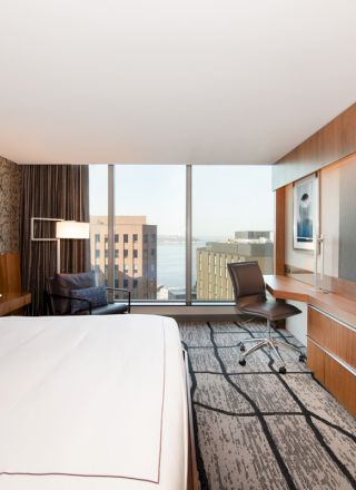The image shows a modern hotel room with a large bed, wall-mounted TV, desk, chair, and a window view of buildings and the water.