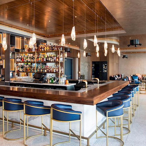 A modern bar with a central counter, blue stools, stylish lighting, and a wide selection of bottles. Cozy seating area in the background.