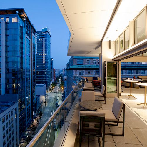 A modern rooftop terrace overlooking a cityscape at dusk, featuring outdoor seating and tables along the edge of a tall building.
