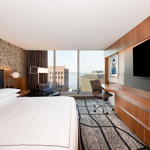 A modern hotel room with a large bed, flat-screen TV, desk, and window view of buildings and water is shown in this image.