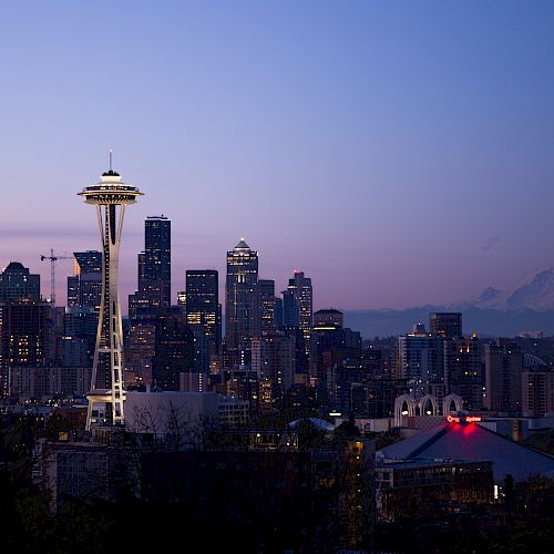 The image shows Seattle's skyline at dusk, with the Space Needle and Mount Rainier in the background, under a clear sky.