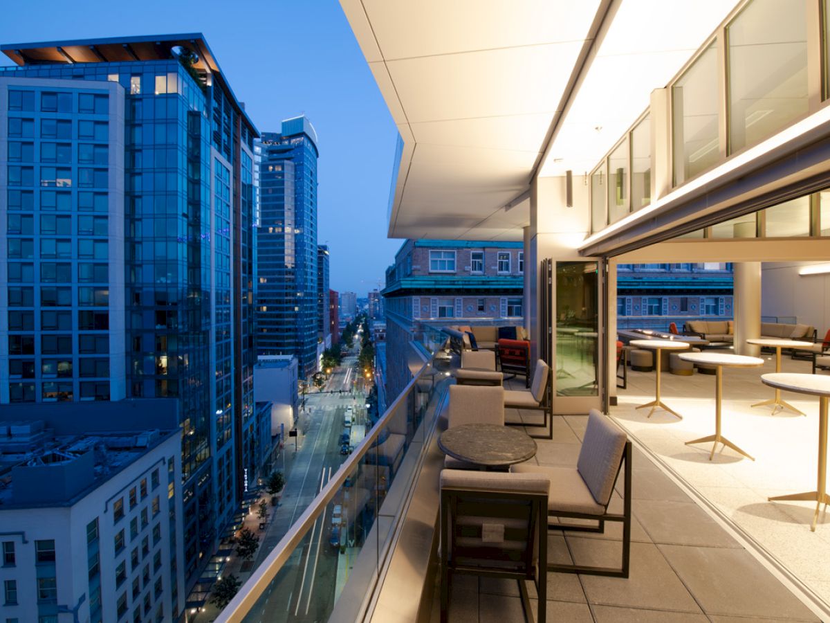 A modern rooftop terrace with tables and chairs overlooks a city street lined with tall buildings during evening.