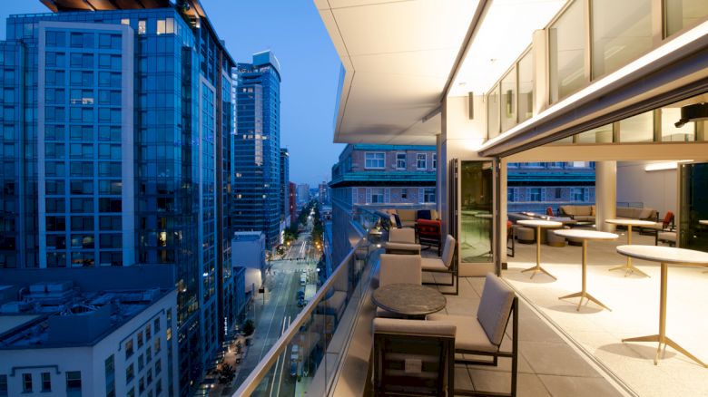 A modern rooftop terrace with tables and chairs overlooks a city street lined with tall buildings during evening.