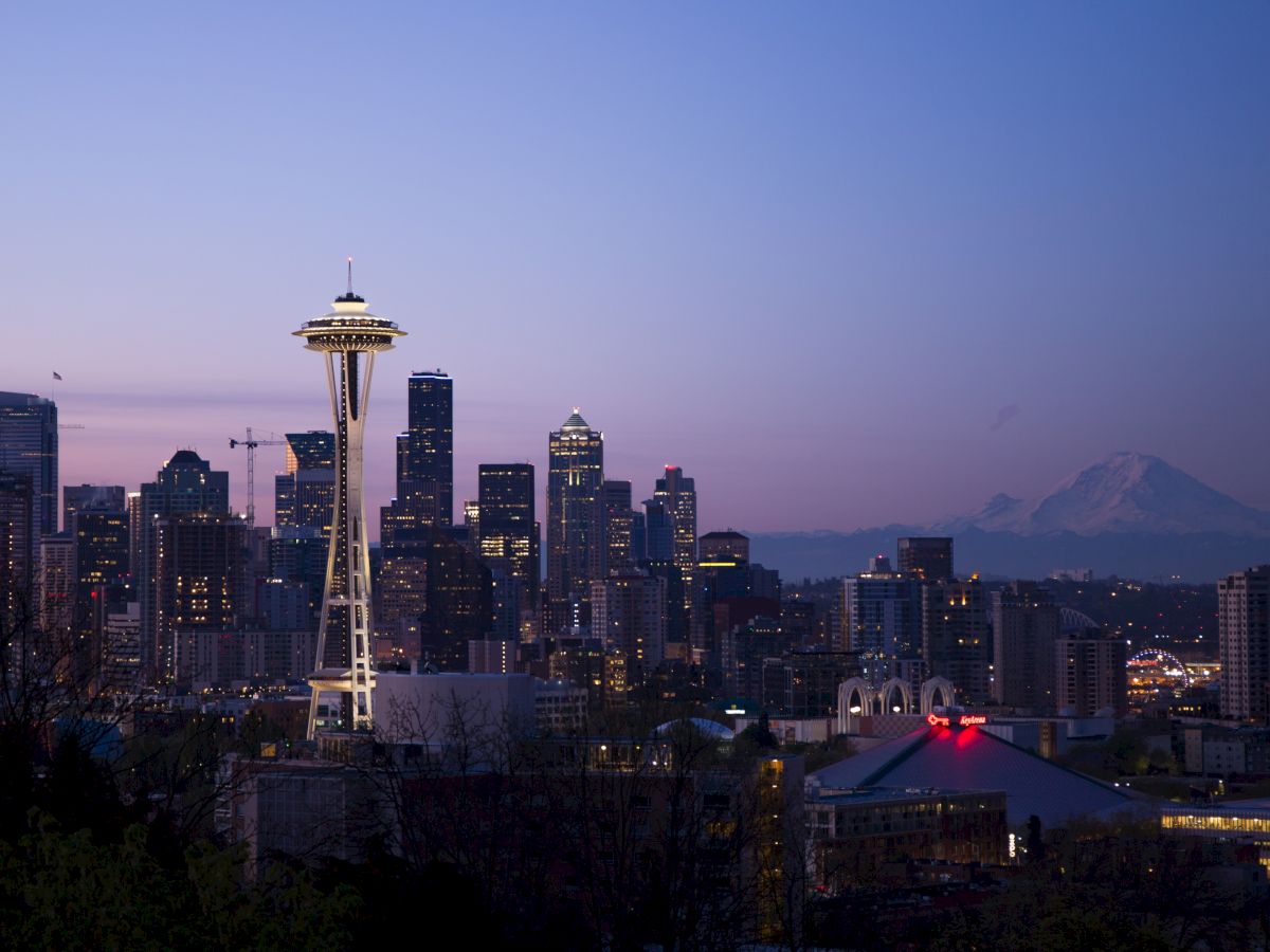 A cityscape of Seattle at dusk with the Space Needle, downtown skyline, and Mount Rainier visible in the background against a colorful sky.