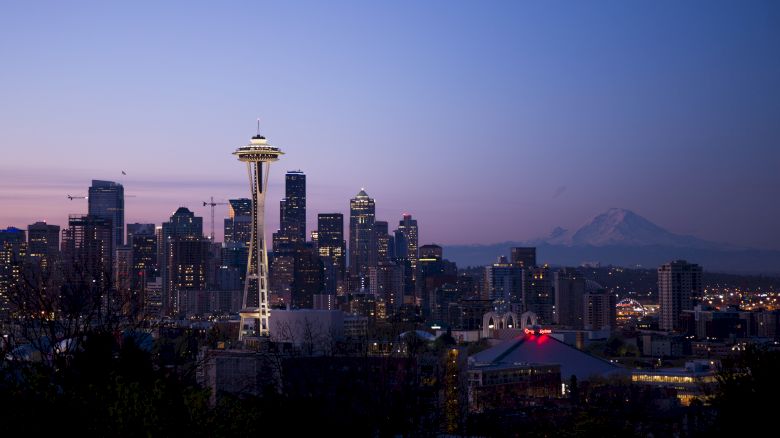 A cityscape of Seattle at dusk with the Space Needle, downtown skyline, and Mount Rainier visible in the background against a colorful sky.