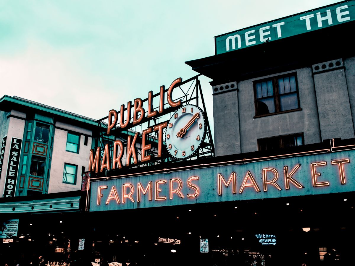 The image shows a neon sign for a Public Market and Farmers Market, with a clock, set against buildings in an urban environment.