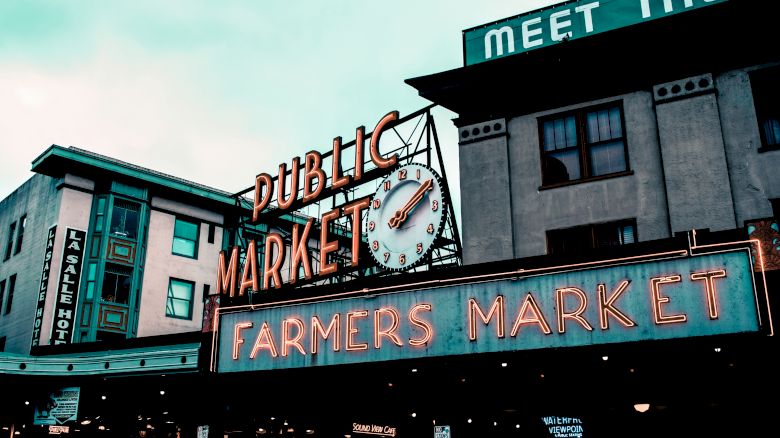 The image shows a neon sign for a Public Market and Farmers Market, with a clock, set against buildings in an urban environment.