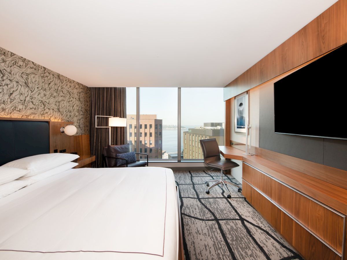 A modern hotel room with a large bed, desk, chair, flat-screen TV, patterned walls, and a view of city buildings and water through the window.