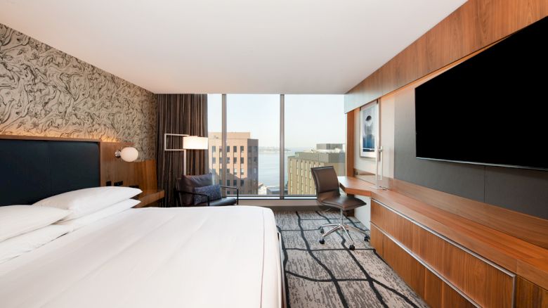 A modern hotel room with a large bed, desk, chair, flat-screen TV, patterned walls, and a view of city buildings and water through the window.