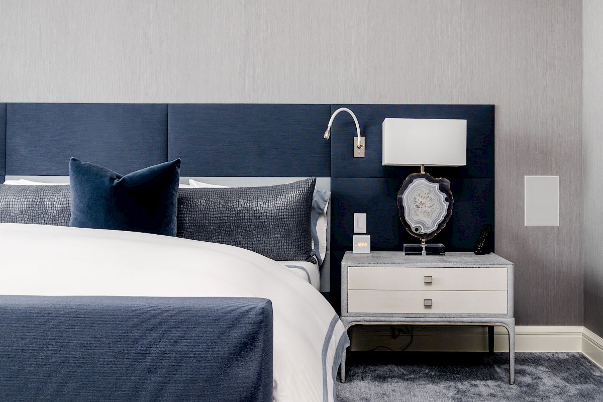 A modern bedroom with a navy and white color scheme features a bed, a headboard, a side table with a lamp, and decorative pillows.