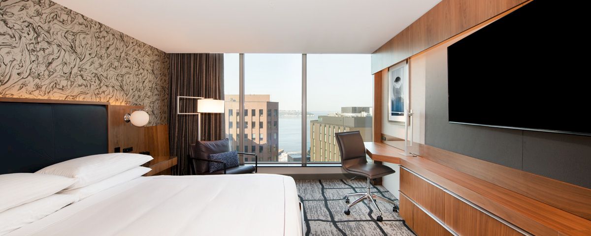 A hotel room with a bed, TV, desk, and chair in front of a window with a city view.