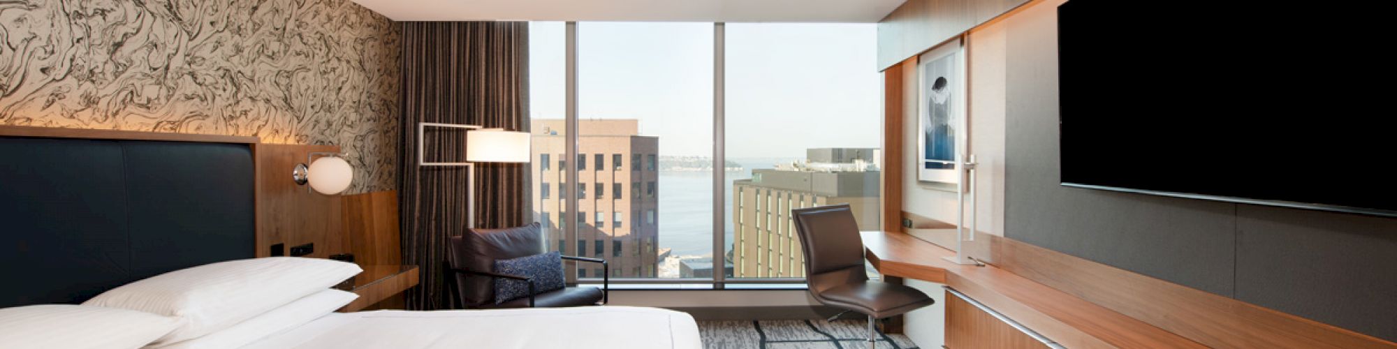 A modern hotel room with a large bed, wall-mounted TV, desk, and chair. The window offers a city view with some buildings visible.